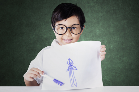 Picture of adorable schoolgirl showing her drawing while sitting in the classroomの写真素材