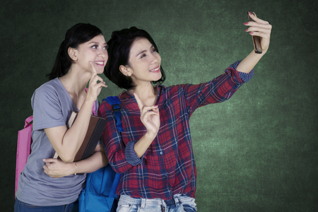 Young female with her friend taking a selfie photo by using a smartphone while standing in the classroomの写真素材