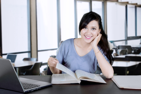 Picture of a beautiful college student smiling at the camera while reading a bookの写真素材