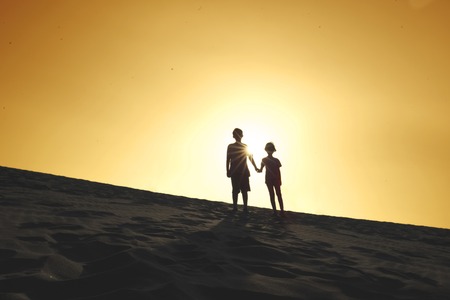 Silhouette of two children holding hands together while standing on the dune at sunset time. Shot on the beachの写真素材
