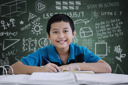 Picture of handsome preteen making notes in the classroom while sitting with scribbles on the chalkboardの写真素材