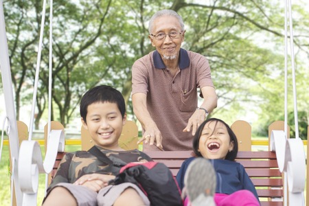 Picture of elderly man pushing his grandchildren on the swing while playing together in the parkの写真素材