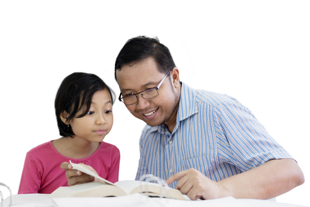 Young father is assisting his daughter to read a book while sitting in the studio, isolated on white backgroundの写真素材