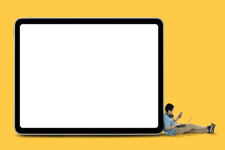 Asian preteen schoolboy using laptop and smartphone while sitting with a blank whiteboard. Shot with yellow backgroundの写真素材
