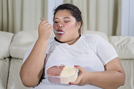 Overweight Asian woman eating ice cream with pleasure at homeの写真素材