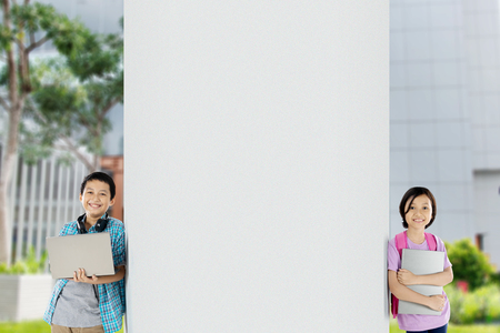 Picture of preteen schoolboy holding a laptop while leaning on a wall with his sister in the schoolの写真素材