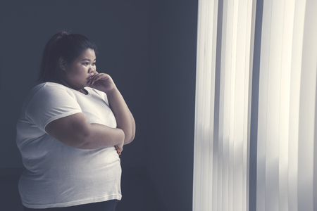 Image of obese woman looks pensive while standing by the windowの写真素材