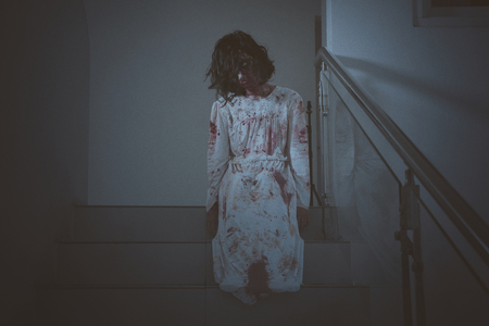 Halloween horror concept. Image of creepy female ghost looking at the camera while sitting in the stairway. Shot at homeの写真素材