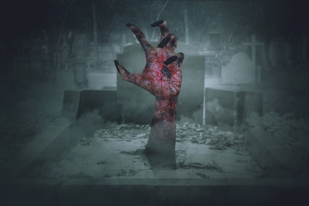 Spooky zombie hand with long nails and bloody wound coming out from graveyard