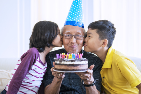 Cheerful old man celebrating his birthday while holding a birthday cake and kissed by grandchildren at homeの写真素材