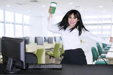 Young businesswoman is dancing near her working table while holding a cup of coffee in the officeの写真素材