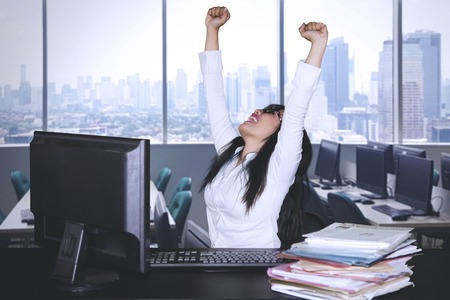 Asian business woman celebrating success by lifting her hands while working with a computerin the officeの写真素材