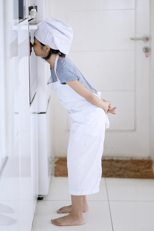Photo of female little chef looking at baked cookie in the oven on the kitchen at homeの写真素材