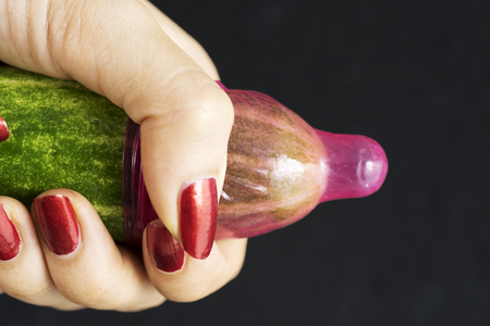 Close up of a young woman holding a cucumber with a condom. Shot with dark backgroundの写真素材