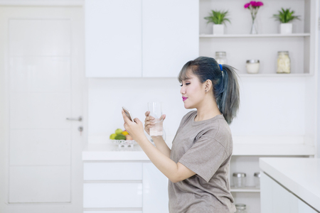 Beautiful woman using a mobile phone while holding a glass of water and standing in the kitchenの写真素材