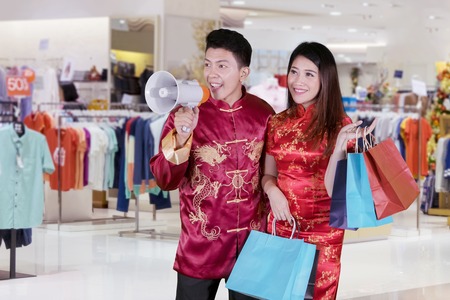 Chinese New Year Concept. Young Chinese couple speaking with a megaphone while carrying shopping bags in the mallの写真素材