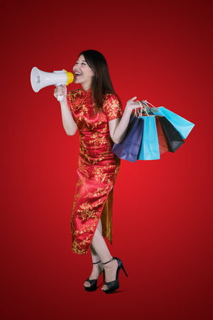 Young Chinese woman speaking with a megaphone while holding shopping bags in the studio. Chinese New Year Conceptの写真素材