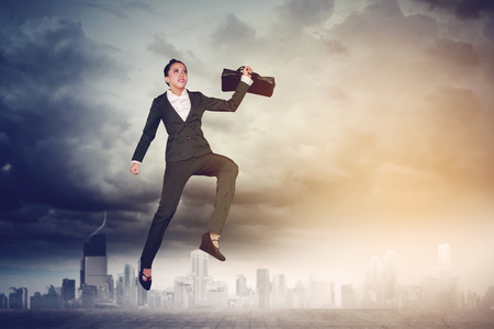 Picture of of young businesswoman carrying a suitcase while jumping on the road with skyscraper backgroundの写真素材