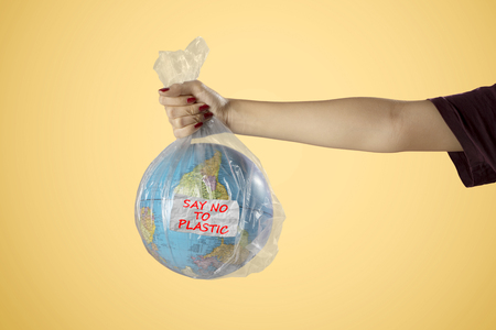 Unknown woman hands holding an earth globe with text of say no to plastic in plastic bag. Shot in yellow backgroundの写真素材