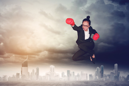 Picture of young female entrepreneur wearing boxing gloves while jumping and punching through at outdoorの写真素材