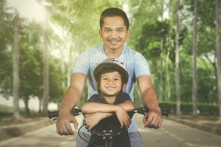 Picture of Asian man looking at the camera while riding bicycle with his son in the parkの写真素材