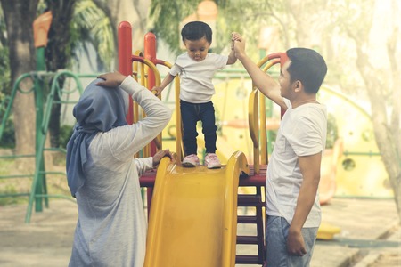 Picture of a baby girl standing on a slide while playing with her parents in the park