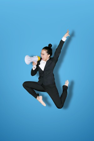 Excited female manager shouting on a megaphone while jumping in the studio with blue backgroundの写真素材