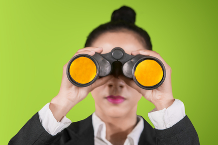 Close up of female manager looking at the camera through binoculars while standing in the studio with green screenの写真素材