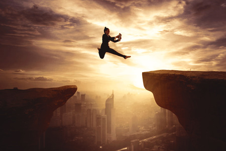Silhouette of young businesswoman using a digital tablet while jumping gap on the cliffの写真素材