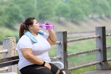 Portrait of overweight woman drinking water after doing run exercises while sitting in the parkの写真素材
