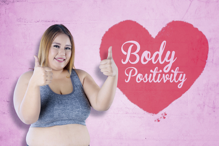 Pretty fat woman smiling at the camera while showing thumbs up near a heart symbol with text of body positivityの写真素材
