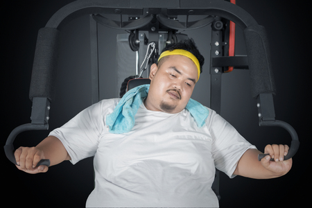 Picture of young fat man looks tired while sitting on the exercise machine. Shot in the dark roomの写真素材