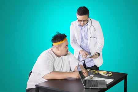 Male doctor showing medical result on the tablet while discussing with his fat patient in the studioの写真素材