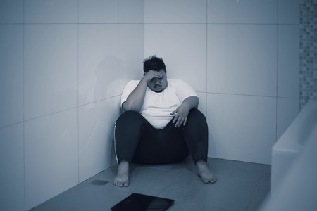 Weight loss failure concept. Young obese man looks sad while sitting with a scale in the bathroomの写真素材