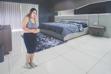 Image of obese woman wearing sportswear while having abdominal pain in the bedroomの写真素材