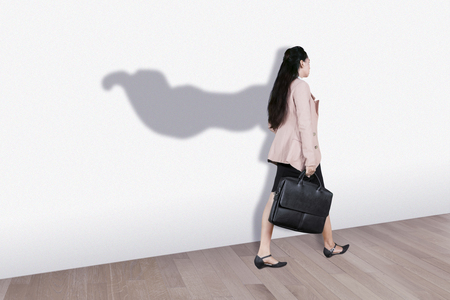 Picture of young businesswoman carrying a suitcase while walking with superhero cape shadow on the wallの写真素材