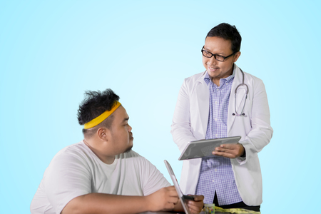 Male physician showing medical result on the tablet while discussing with his fat patient in the studioの写真素材