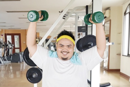 Happy fat man lifting two dumbbells while exercising in the gym centerの写真素材