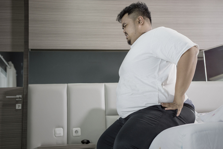 Overweight man having backache while sitting on the bed. Shot at homeの写真素材