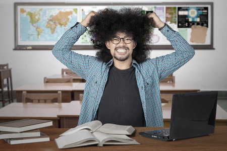 Stressful afro college student pulling his hair in the class with books and laptop on the table