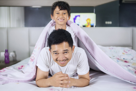 Happy young man and his son cuddling on the bed while smiling and looking at the cameraの写真素材