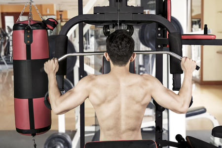 Back view of young man using fitness machine for trains his muscular arms at the gym
