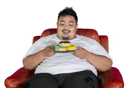 Happy fat man holding a plate of donuts while sitting on the armchair, isolated on white backgroundの写真素材