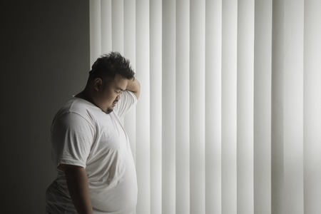 Picture of overweight man looks lonely while standing near the window in the dark roomの写真素材