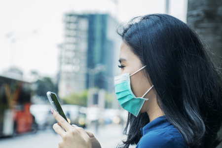 Side view of young woman wearing a mask while using a smartphone at outdoors. Air Pollution Conceptの写真素材