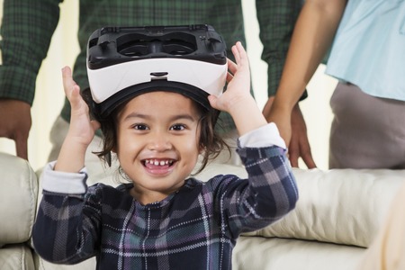 Close up of little girl smiling at the camera while holding a virtual reality goggles and sitting on...