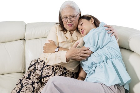Picture of young sad woman embracing her mother while sitting on the couch, isolated on white backgroundの写真素材