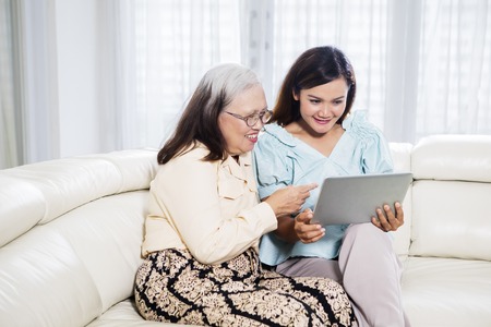 Picture of old woman using a digital tablet with her daughter while sitting together on the couch