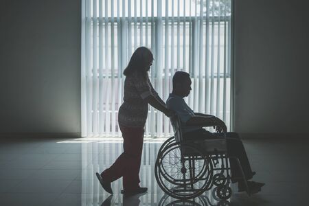 Silhouette of aged woman pushing her husband on a wheelchair after recovering from stroke disease