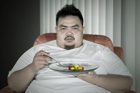 Close up of Obese man looks unhappy while eating a plate of healthy food on the sofa and sitting at homeの写真素材